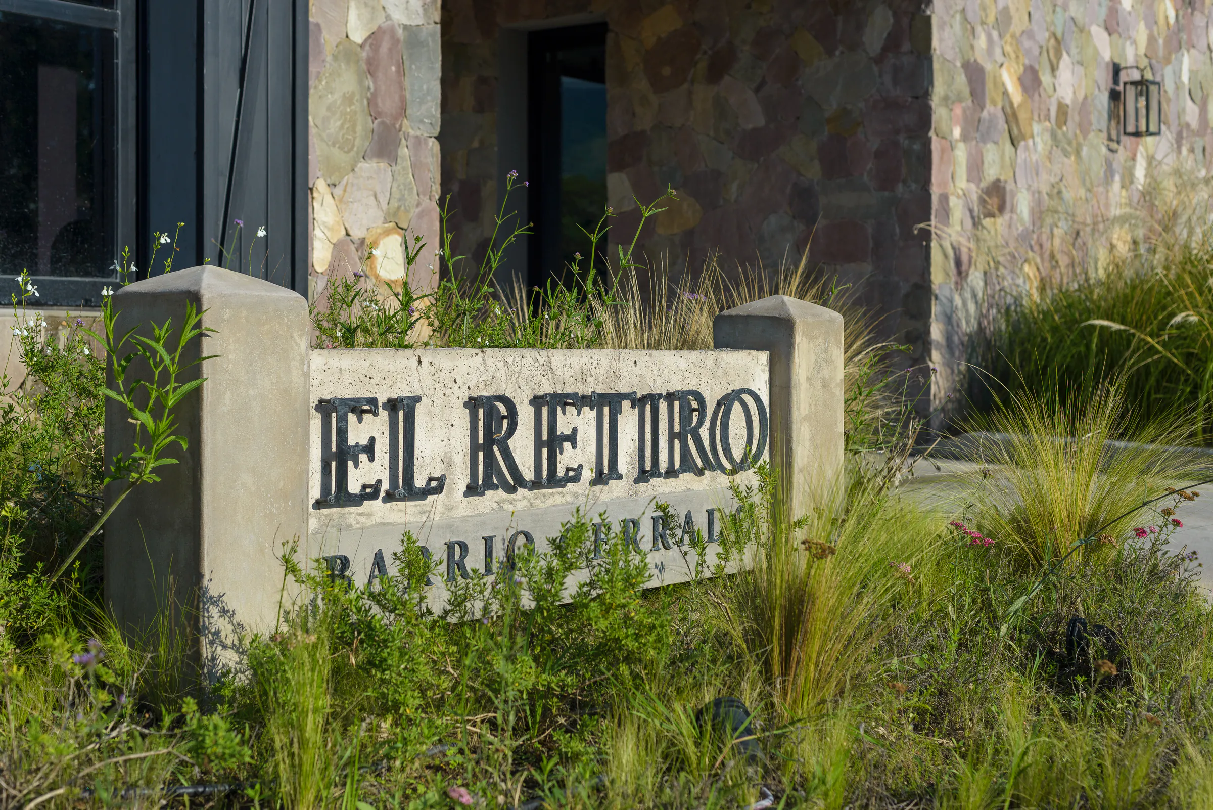 Cartel de hormigón con las letras de El Retiro, barrio cerrado, entre gramíneas y flores, delante de un muro de piedra