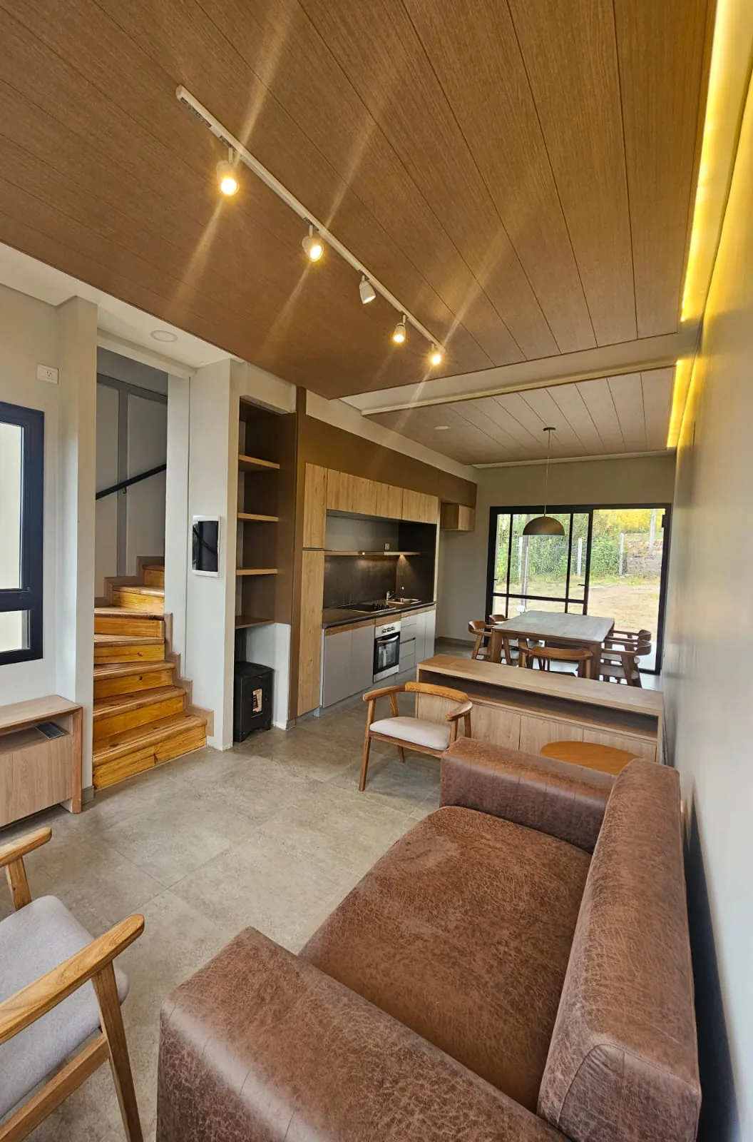 Interior de una casa Ele Haus terminada: estar con un sillón marrón y sillones de madera con almohadones grises, escalera de madera, salamandra negra y cocina con muebles de madera clara, bajo un cielorraso de madera con un riel de spots.