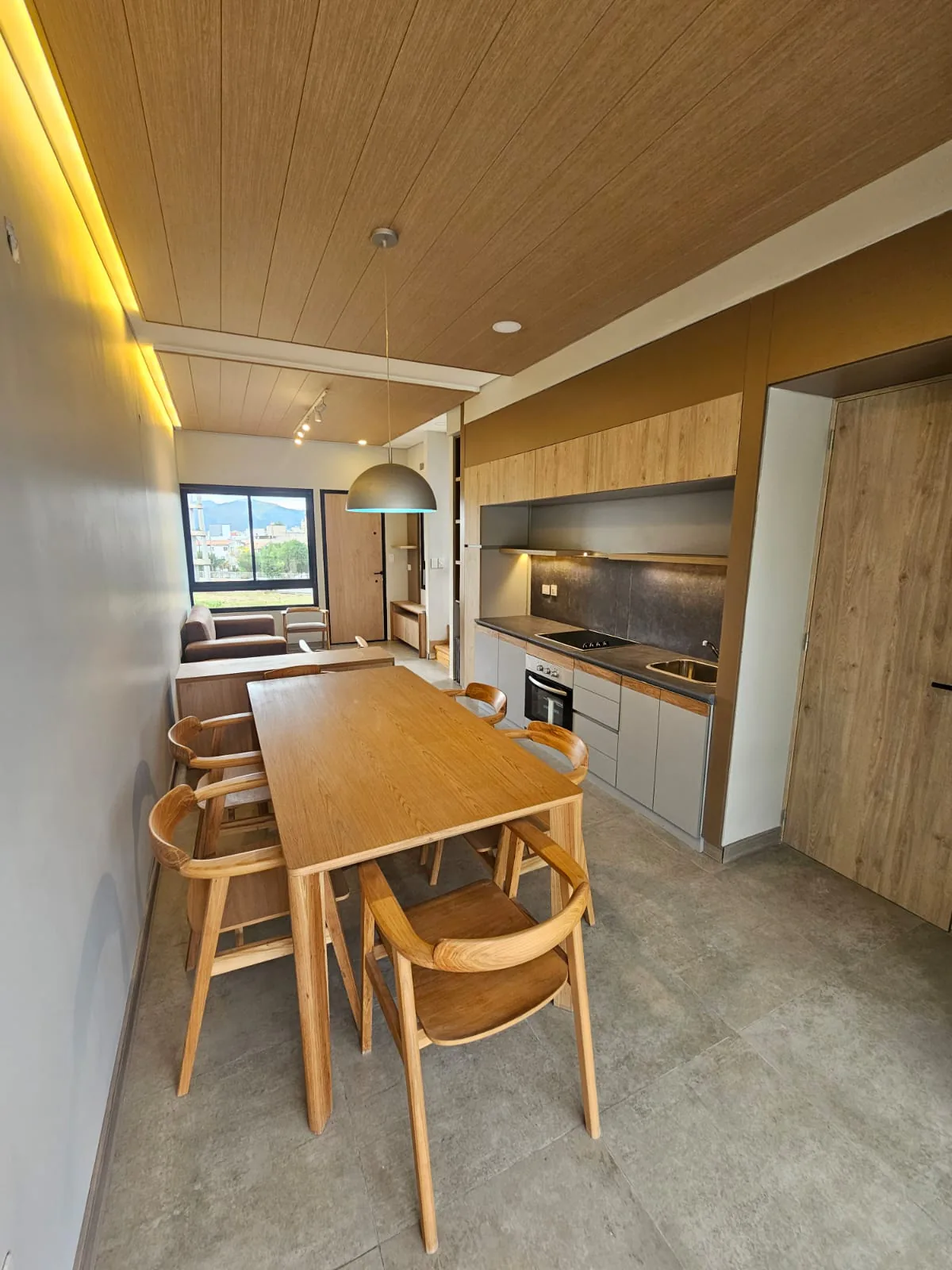 Interior de una casa Ele Haus terminada: mesa de madera con seis sillas junto a la cocina gris y madera, lámpara colgante gris y, al fondo, el estar con una ventana que da a los cerros.