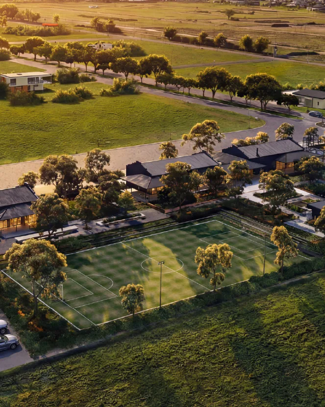 Render aéreo al atardecer del sector del club: edificios de techos oscuros entre árboles, una cancha de fútbol 7, dos canchas de tenis azules y un estacionamiento con autos, con campos y casas detrás.